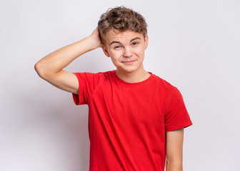 Wall Mural - Portrait of pensive teen boy on grey background. Thoughtful teenager holding hand near head, looking at camera. Handsome caucasian funny child student in red t-shirt.