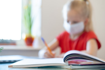 Wall Mural - Blurred schoolgirl in medical mask studying at home, doing school homework, writing in notebook. Reading training books on table.coronavirus quarantine. Distance learning online education