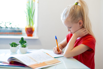 Wall Mural - Serios blonde schoolgirl sitting at home and doing school homework, writing in notebook. Training books on table. Back to school. Distance learning online education