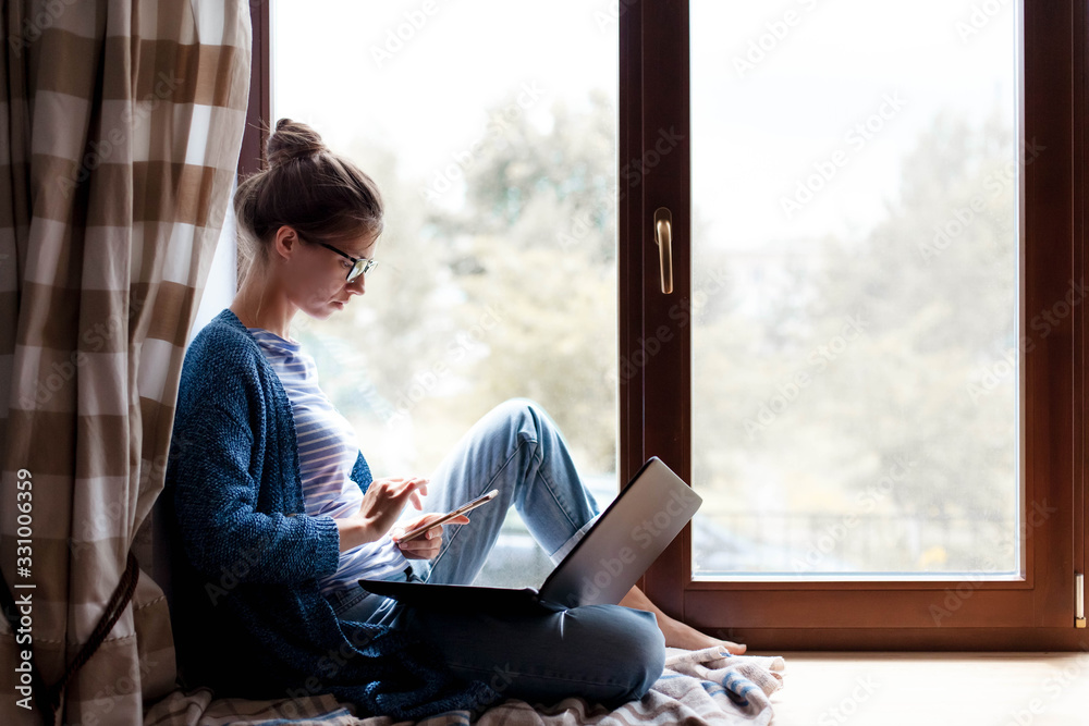 Young woman sitting in window with laptop and smartphone - obrazy, fototapety, plakaty 