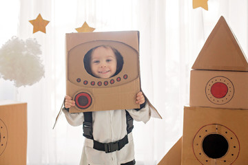 Sweet toddler boy, dressed as an astronaut, playing at home with cardboard rocket and handmade helmet