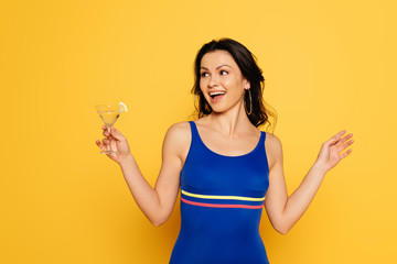 Wall Mural - cheerful woman in blue swimsuit holding glass of cocktail and looking away isolated on yellow