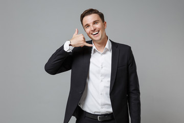 Cheerful young business man in classic black suit shirt posing isolated on grey background. Achievement career wealth business concept. Mock up copy space. Doing phone gesture like says call me back.