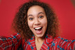 © ViDi Studio - Close up of excited young african american girl in pajamas homewear rest at home isolated on red background. Relax good mood lifestyle concept. Mock up copy space. Doing selfie shot on mobile phone.