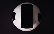 Live Football Betting Concept, Online Broadcast, Blank Smartphone Display On Classic Soccer Ball Isolated On Black Background. Sports Gambling Copy Space App, Play Entertaining Games On Mobile Phone.