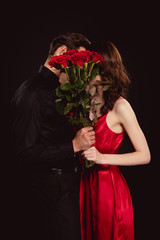 Wall Mural - Side view of elegant couple covering faces with bouquet of red roses isolated on black