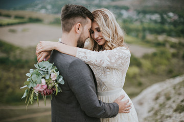 Smiling bride and groom spending time together. Posing on the mountain hills background. Dressed in white dress beautiful blonde caucasian bride and handsome groom. Hugs, kissess and enjoy the company