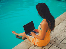 Remote Online Working Digital Nomad Women From The Side With Long Hair And Laptop Sitting At A Sunny Blue Water Pool 