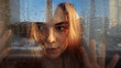 The girl looks suspiciously behind the glass. There are raindrops on the glass. The girl has blond hair and yellow eyes. Hands are pressed to the glass. Outside the window a blue sky.