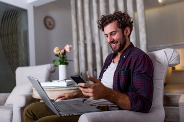 Wall Mural - Young handsome man making video call with friends while sitting on sofa at his modern home.Concept of happy business people. Business man at home checking statistics on mobile phone and laptop