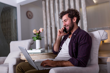 Young attractive smiling guy is browsing at his laptop and talking oh mobile phone, sitting at home on the cozy sofa, wearing casual outfit. Freelance business work from home concept