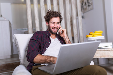 Wall Mural - Young attractive smiling guy is browsing at his laptop and talking oh mobile phone, sitting at home on the cozy sofa, wearing casual outfit. Freelance business work from home concept