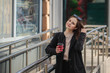 © Юлия Буракова - cheerful woman on a city street drinks coffee on a sunny day and looks at the camera. coffee to go. Portrait of a girl with a drink in hands