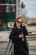 © Юлия Буракова - cheerful woman on a city street drinks coffee in a sunny day in sunglasses and looks away. coffee to go. Portrait of a girl with a drink in hands, vertical photo