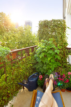 Home Office In Summer: Woman's Feet In Flip-flops On A Footrest On A City Balcony With Lots Of Plants.