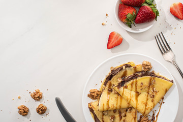 Wall Mural - top view of tasty crepes with chocolate spread and walnuts on plate near bowl with strawberries and cutlery on grey background