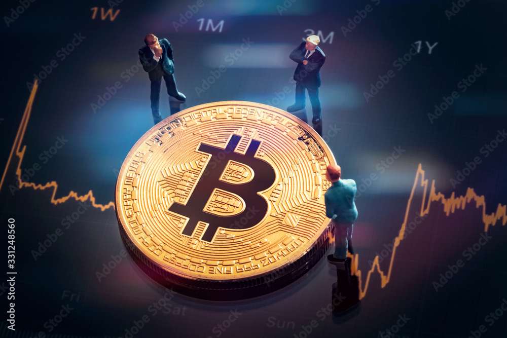 Business meeting about the investment decision for bitcoin: Macro miniature businessman figurines standing near the shiny golden bitcoin. Colorful stock market graphic chart on background. 