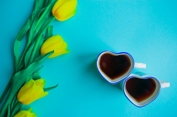 Happy spring card. Creative greeting card with the bouquet of beautiful spring yellow tulips flowers on the light blue background and two cups of tea. Copy space. Flat lay, still life, top view.