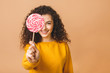 © denis_vermenko - Surprised curly girl eating lollipop. Beauty Model woman holding pink sweet colorful lollipop candy, isolated on beige background.