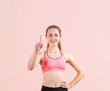 © A Stock Studio - Young fitness woman points a finger in upward standing on a light pink background. Sportswoman pointing up on copy space and looking at the camera.