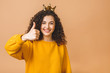 © denis_vermenko - Gorgeous beautiful girl with curly brown hair and wearing casual and holding crown on head isolated over beige studio background. Thumbs up.