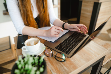 Closeup photo. Student girl sitting with laptop and papers in cafe with cup of coffee and studying. Lady freelancer works on the laptop and drinks coffee in a coffee shop.Working in a cafe. Background