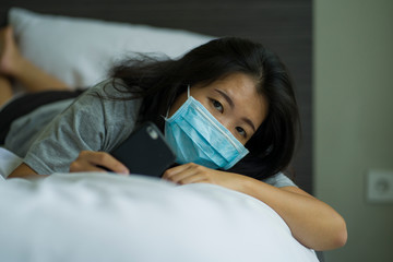 virus outbreak - young beautiful sad and worried Asian Chinese woman wearing medical face mask in quarantine and lockdown at home in panic as prevention vs coronavirus