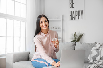 Canvas Print - Beautiful young woman drinking water while using laptop at home