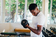 © twinsterphoto - Young African American man standing and lifting a dumbbell with the rack at gym. Male weight training person doing a biceps curl in fitness center