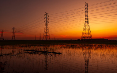 High voltage electric pylon and electrical wire with sunset sky. Electricity poles. Power and energy concept. High voltage grid tower with wire cable at rice farm field near industrial factory.