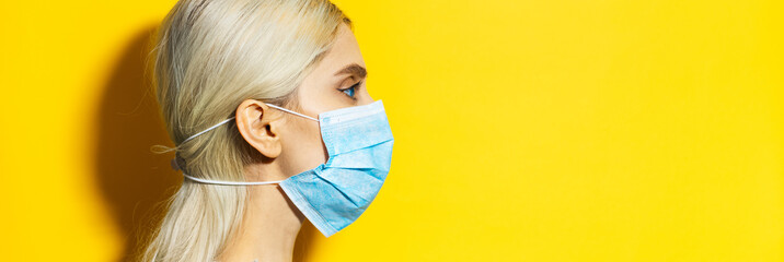 Wall Mural - Studio profile portrait of young blonde girl with blue eyes, wearing medical flu mask, small protection from coronavirus. Isolated on yellow background with copy space.