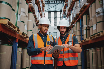 Wall Mural - Focused manager and employee standing in warehouse discussing inventory stock while standing between cardboard packed goods with safety hardhat and vest