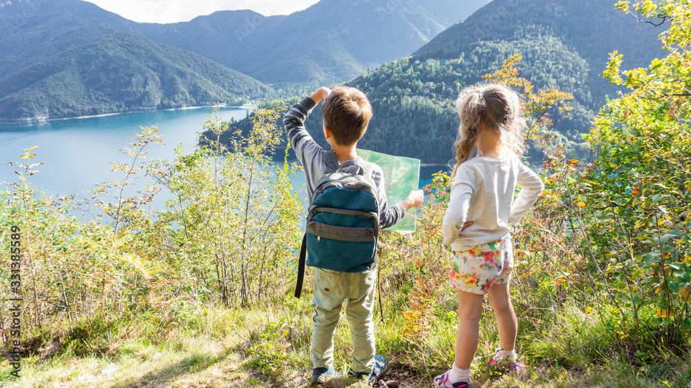 Two scout children stand on a tourist route along a camping road on the shore of a mountain lake Ledro in the Alps. Kids hiking in the forest in Alps mountains - obrazy, fototapety, plakaty 