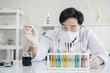 © mrwinn - A male scientist with black hair wearing white coat and protective glassware and face mask dropping liquid solution from a pipet into a test tube looking serious in a laboratory setting.