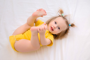 Wall Mural - Toddler girl in yellow bodysuit smiles, lies in bed at home