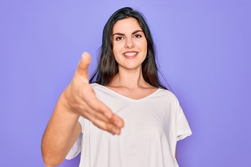 Poster - Young beautiful brunette woman wearing casual white t-shirt over purple background smiling friendly offering handshake as greeting and welcoming. Successful business.