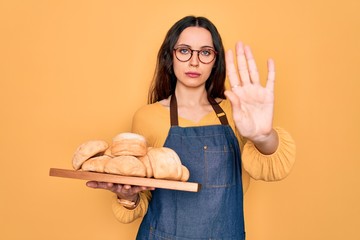 Wall Mural - Young beautiful baker woman with blue eyes wearing apron holding tray with bread with open hand doing stop sign with serious and confident expression, defense gesture