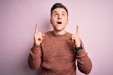 Young handsome caucasian man wearing casual winter sweater over pink isolated background amazed and surprised looking up and pointing with fingers and raised arms.