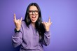 © Krakenimages.com - Young brunette woman wearing glasses over purple isolated background shouting with crazy expression doing rock symbol with hands up. Music star. Heavy concept.