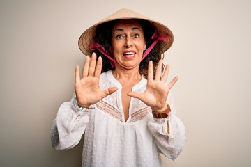 Canvas Print - Middle age brunette woman wearing asian traditional conical hat over white background afraid and terrified with fear expression stop gesture with hands, shouting in shock. Panic concept.