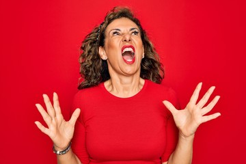 Middle age senior brunette woman wearing casual t-shirt standing over red background crazy and mad shouting and yelling with aggressive expression and arms raised. Frustration concept.