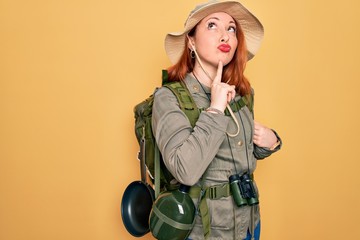 Poster - Young redhead backpacker woman hiking wearing backpack and hat over yellow background Thinking concentrated about doubt with finger on chin and looking up wondering