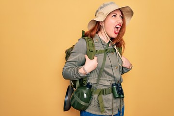 Poster - Young redhead backpacker woman hiking wearing backpack and hat over yellow background angry and mad screaming frustrated and furious, shouting with anger. Rage and aggressive concept.