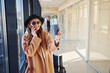 © standret - Young woman in sunglasses, warm clothes and hat standing indoors in airport with phone and tickets in hands