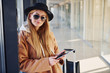 © standret - Young woman in sunglasses, warm clothes and hat standing indoors in airport with phone and tickets in hands