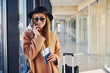 © standret - Young woman in sunglasses, warm clothes and hat standing indoors in airport with phone and tickets in hands