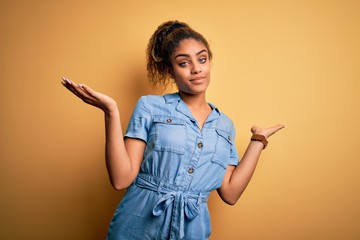 Sticker - Young beautiful african american girl wearing denim dress standing over yellow background clueless and confused expression with arms and hands raised. Doubt concept.