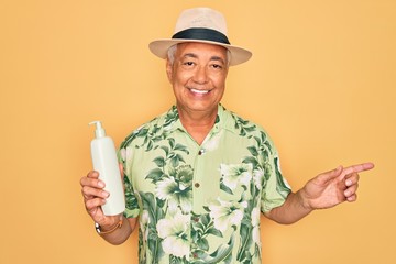 Canvas Print - Middle age senior grey-haired man wearing summer hat holding sun protection cream very happy pointing with hand and finger to the side