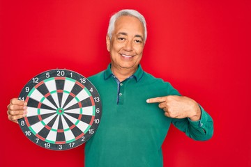 Wall Mural - Middle age senior grey-haired man holding competition dartboard target over red background with surprise face pointing finger to himself