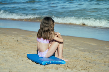 a girl seen from the side on the beach by the sea sitting on a blue surfboard